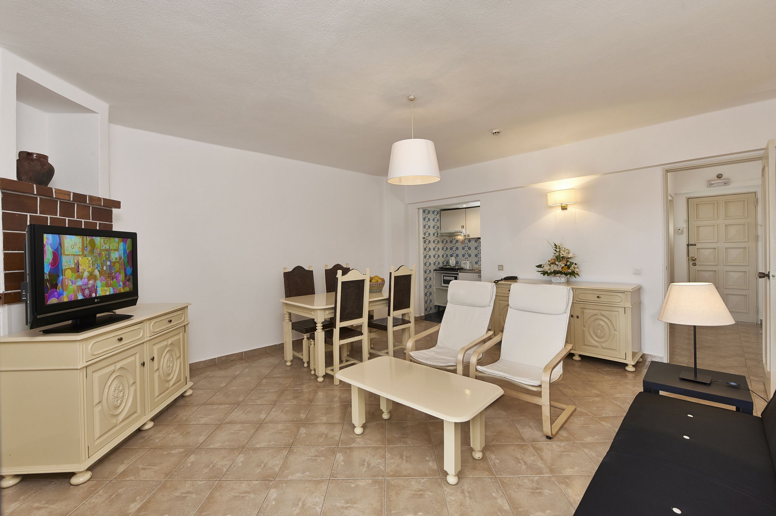 A living room with a television, a dining table with chairs, a bench, a lamp, and a fireplace.