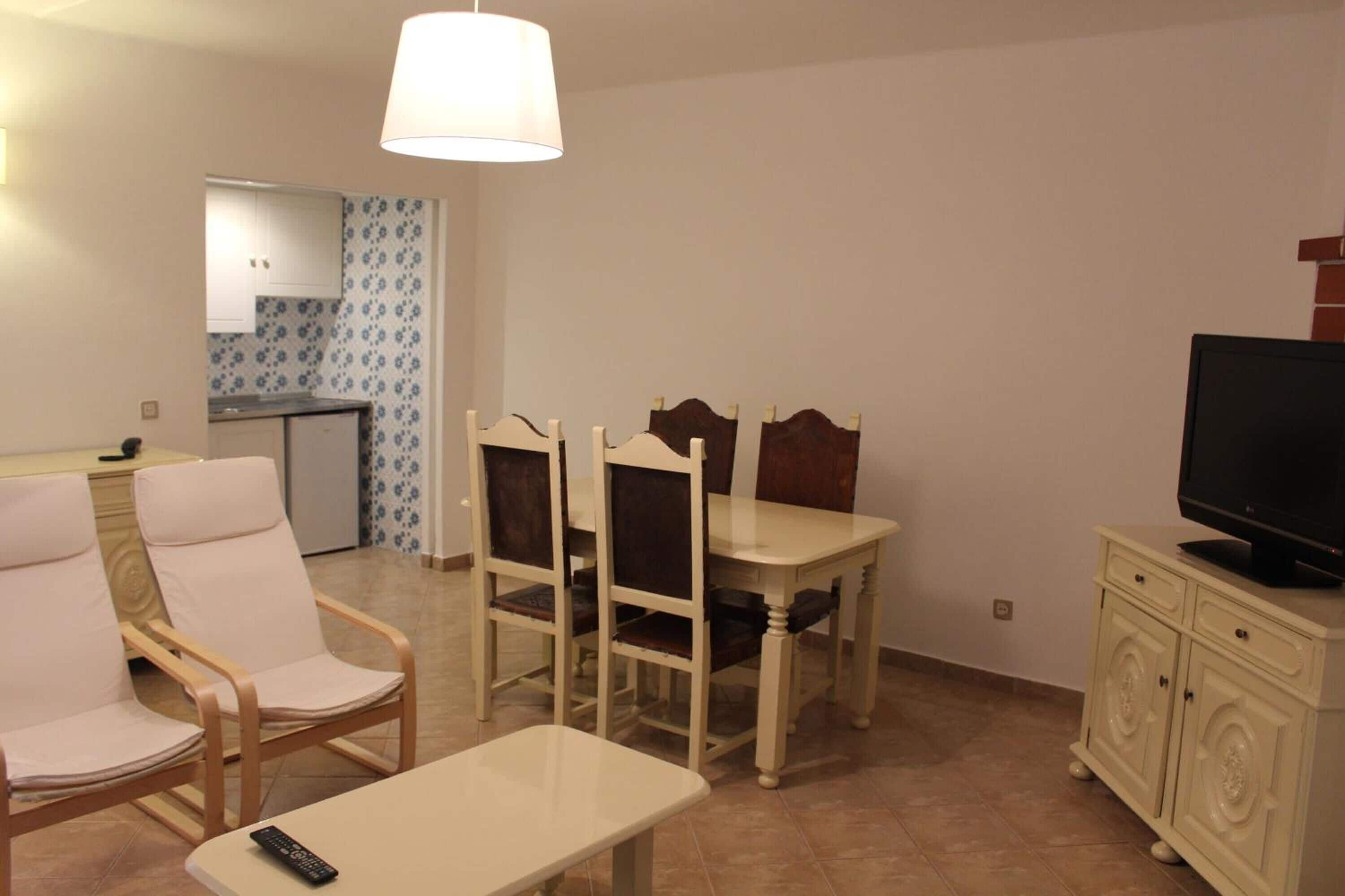 Un espacio habitable pequeño con una mesa de comedor, dos sillas, un televisor sobre un mueble y una cocina al fondo.
