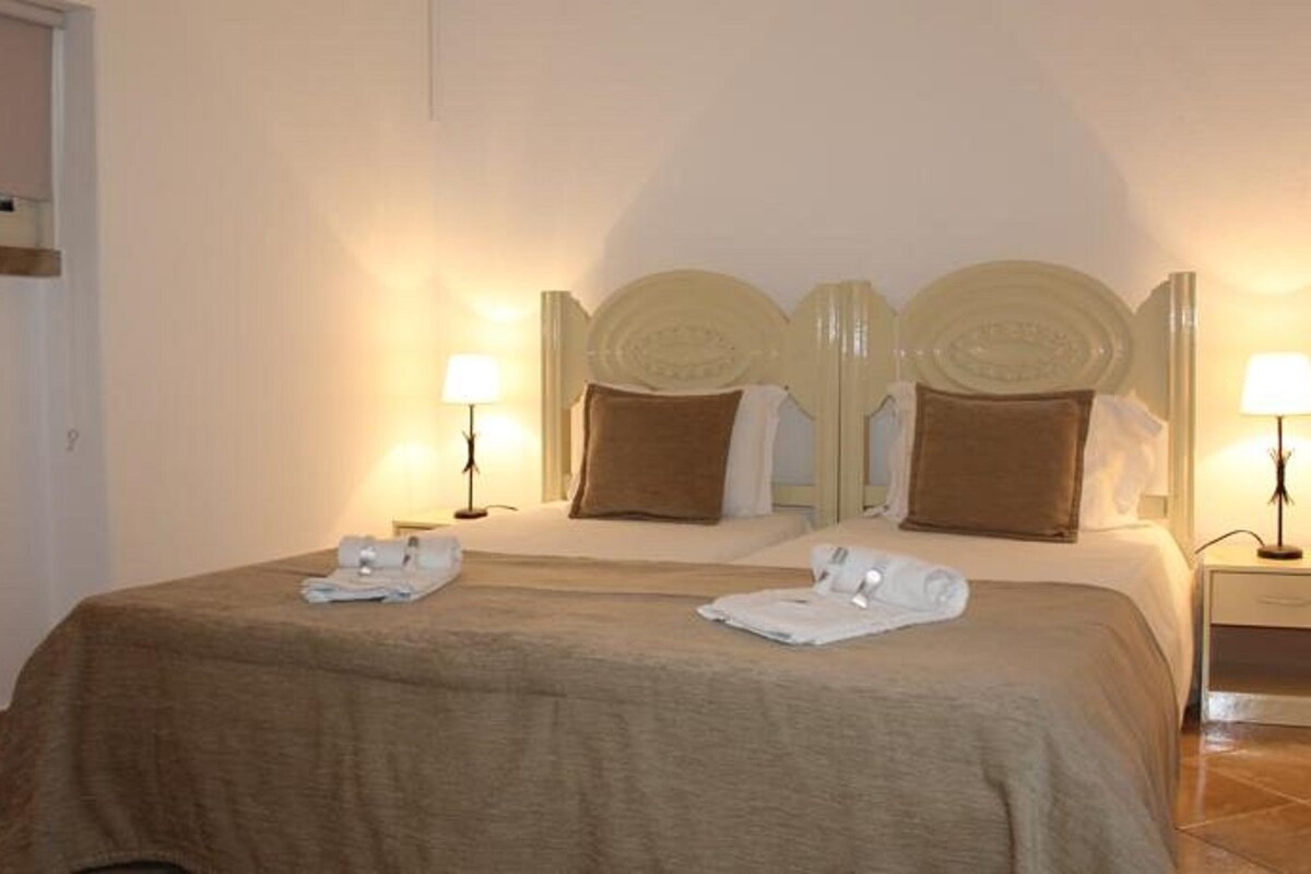 Una habitación de hotel con una cama grande, dos mesitas de noche con lámparas y una puerta a la izquierda.