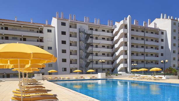 A swimming pool with sun loungers and umbrellas, surrounded by a multi-story hotel building with balconies.