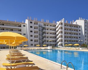 Une piscine dotée de chaises longues et de parasols, entourée d’un immeuble d’hôtel à plusieurs étages, avec des balcons.