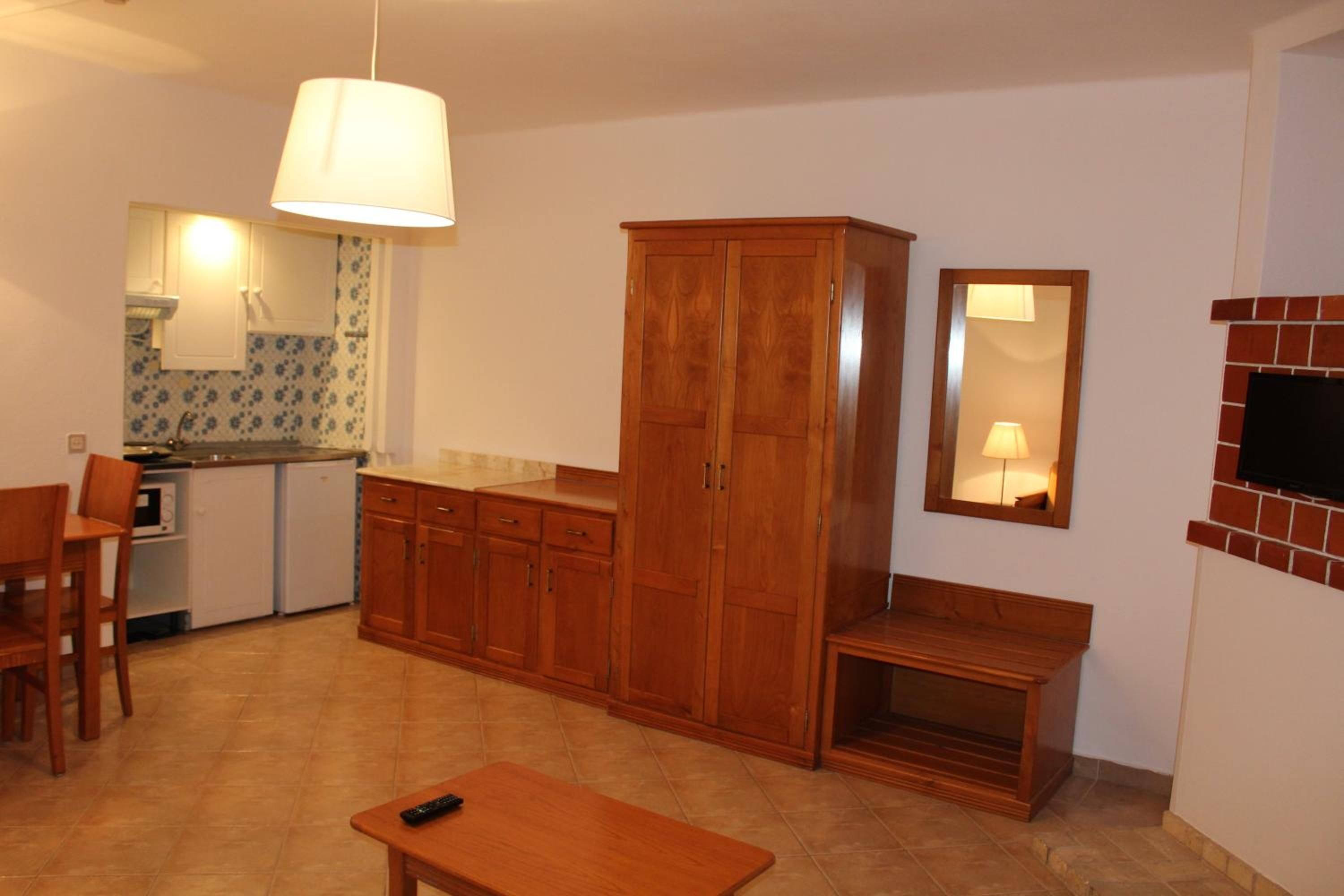 Una cocina compacta con armarios de madera, un comedor con mesa y sillas, y un pequeño rincón de estar con un banco.