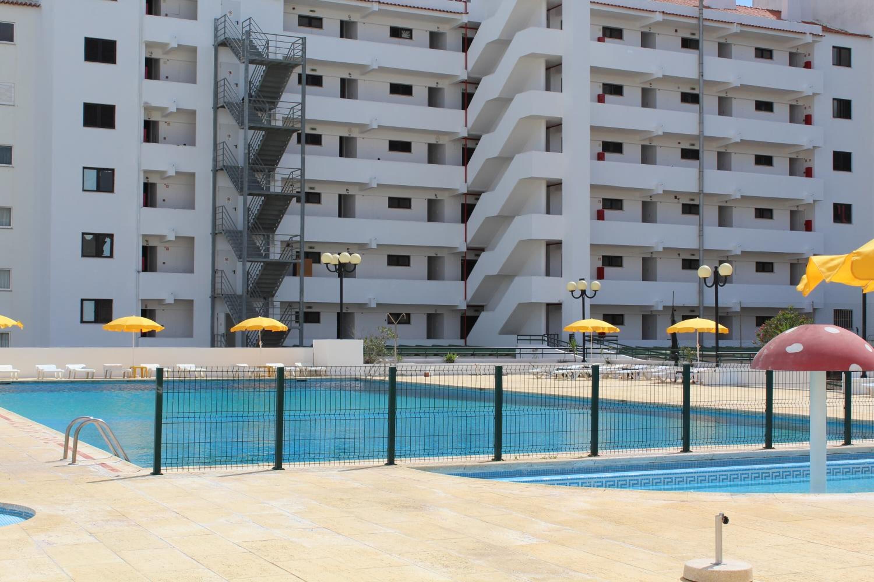 A swimming pool with a net fence, yellow umbrellas, and a building with multiple balconies in the background.