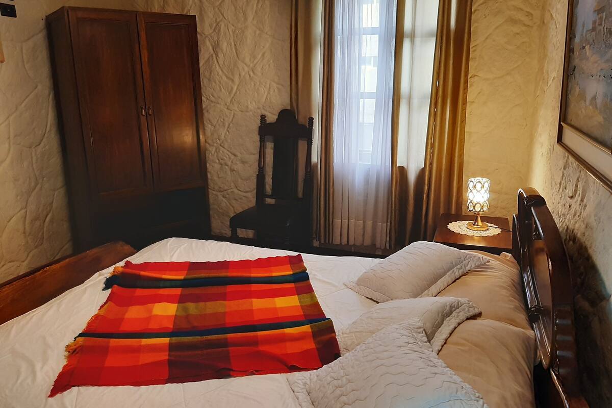 A bedroom with a bed, a wooden wardrobe, a chair, a nightstand with a lamp, and a window with curtains.