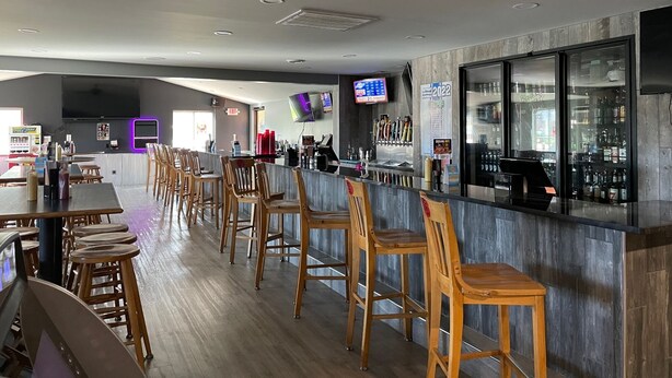 Un área de bar moderna con taburetes de madera, una barra con pantalla digital y un refrigerador lleno de bebidas.