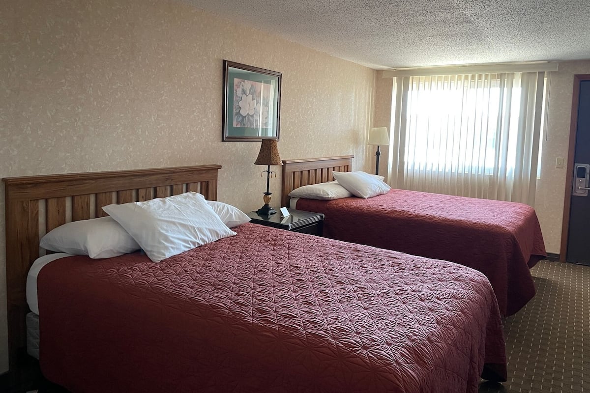 Quarto de hotel com duas camas, cada uma com cabeceira de madeira e roupa de cama vermelha. Há uma moldura na parede, uma luminária na mesa de cabeceira e uma janela com cortinas.