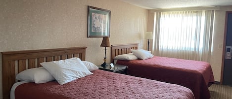 A hotel room with two beds, each with a wooden headboard and red bedding. There is a framed picture on the wall, a lamp on the nightstand, and a window with curtains.