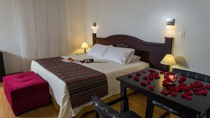 Hotellihuone, jossa on sänky, kaksi yöpöytää, kirjoituspöytä, jonka päällä on ruusun terälehtiä, ja punainen rahi.