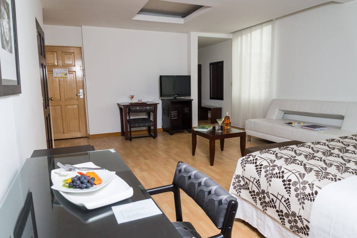 Et hotelværelse med seng, fjernsyn, spisebord med frugt, sofa og et lille bord med en flaske.