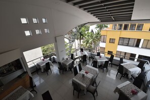 A modern restaurant with multiple tables and chairs, a kitchen area, and a view of the surrounding buildings and greenery.