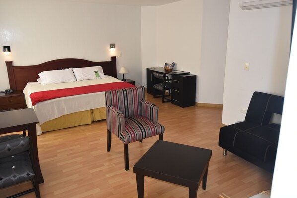 A hotel room with a bed, a striped armchair, a black sofa, a small table, and a desk with a lamp.