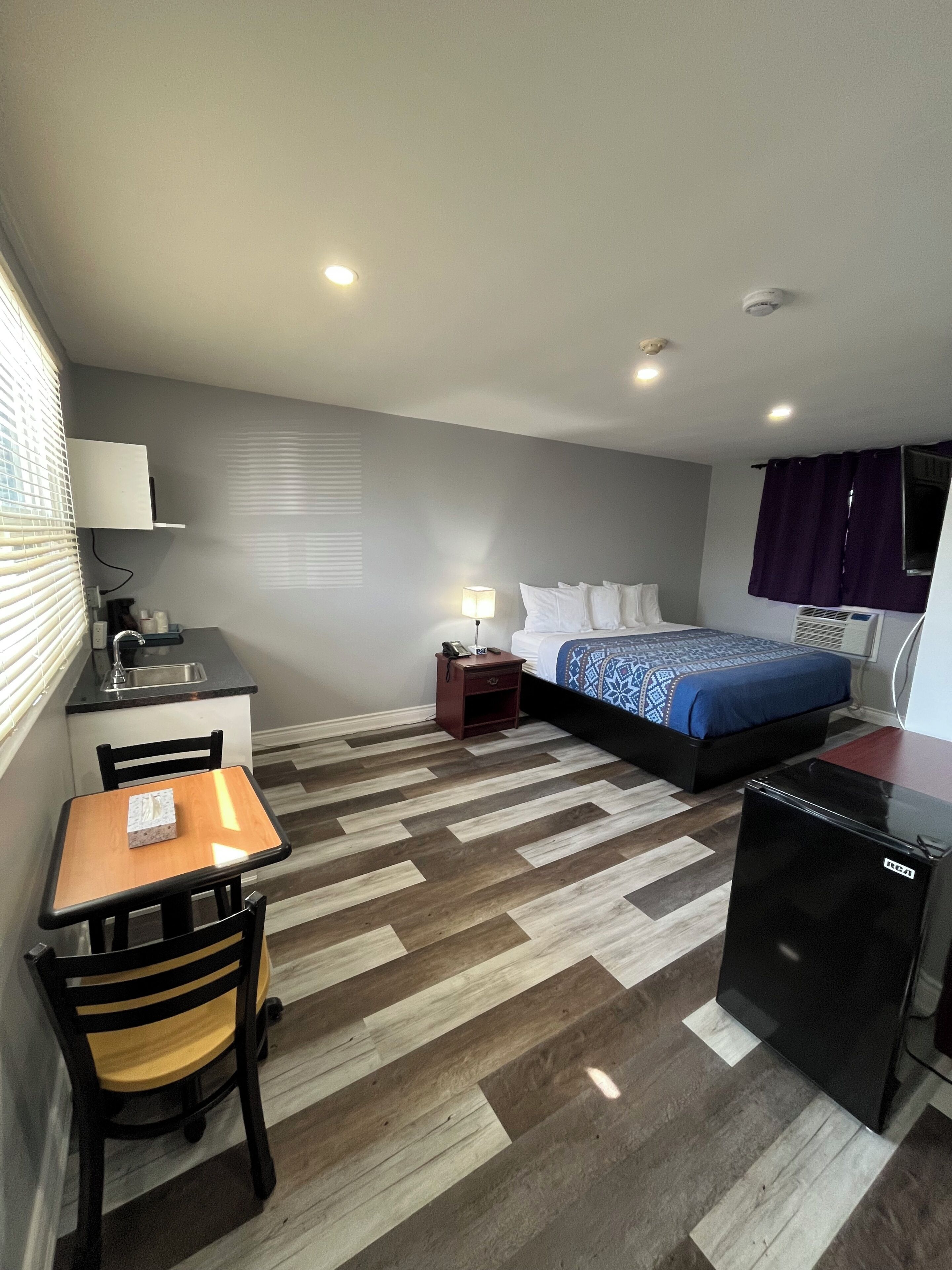 A compact hotel room with a kitchenette, a bed with a patterned bedspread, a small table with a chair, and a refrigerator.