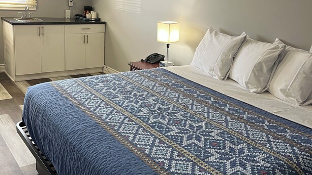 A bedroom with a bed, a nightstand with a lamp, a small desk with a microwave, and a window with blinds. - Cobourg Inn (Hamilton Township)