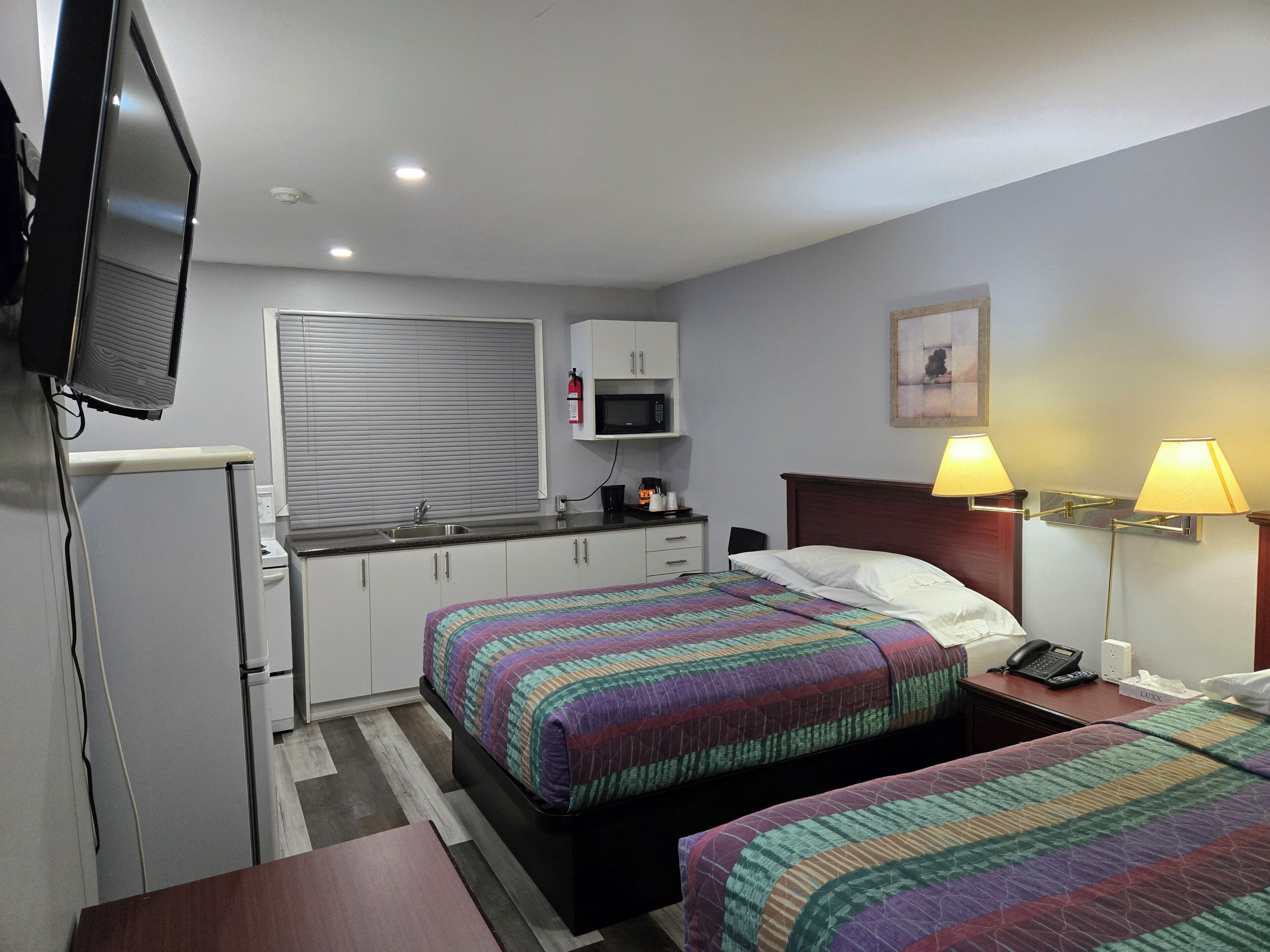 A hotel room with two beds, a kitchenette, a television, and a painting on the wall.