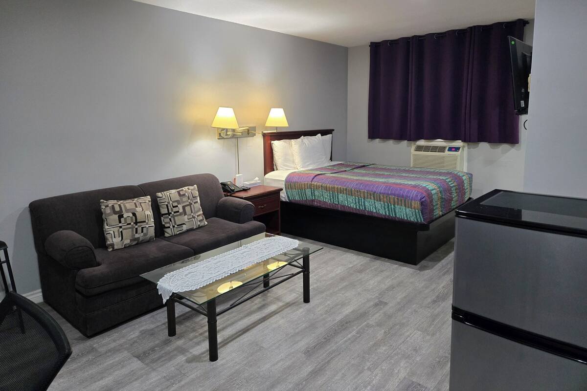 Ein Hotelzimmer mit einem Bett, einem Sofa, einem kleinen Tisch mit weißer Spitzenleinentischdecke, einem Nachttisch mit Lampe und einem Kühlschrank.