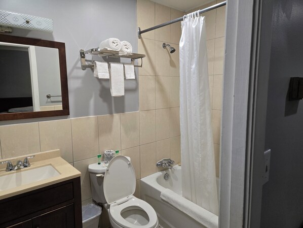 Un baño con inodoro, lavamanos y bañera. Se observan toallas y una cortina de ducha en un perchero.