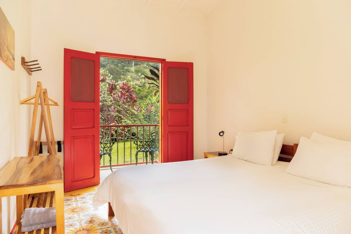 Un dormitorio con una cama, un caballete de madera, una puerta roja y vistas al verdor exterior.