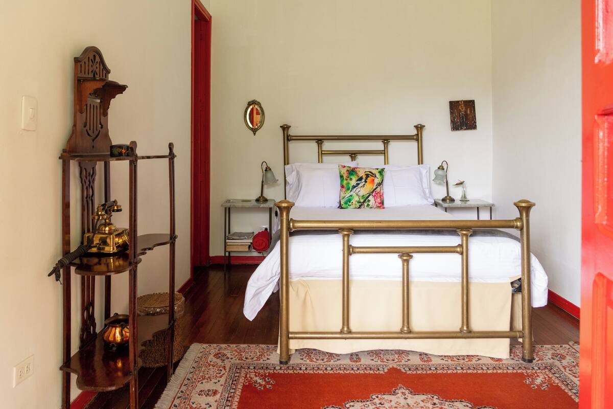Una habitación con una cama, una mesita de noche con lámpara, un estante con teléfono y una puerta roja.