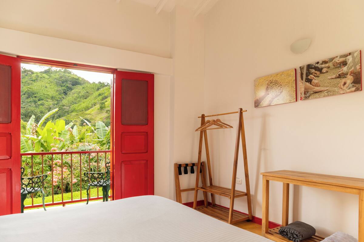 Una habitación con una cama, una mesa de madera, un perchero y un balcón con una puerta roja y vistas al verdor.