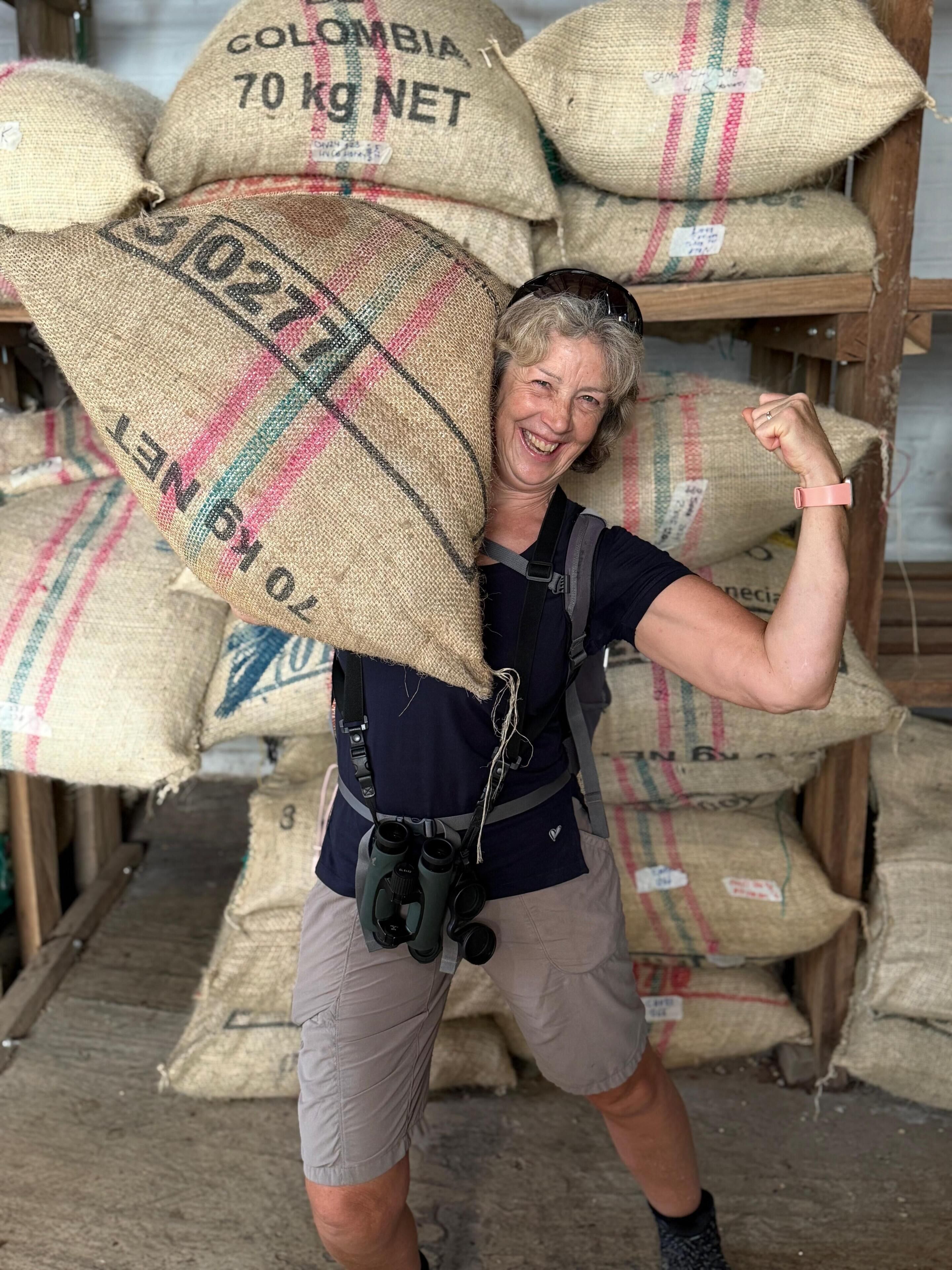 Une personne transportant un grand sac étiqueté « Colombie, 70 kg NET » dans un entrepôt, avec plusieurs autres sacs empilés en arrière-plan.