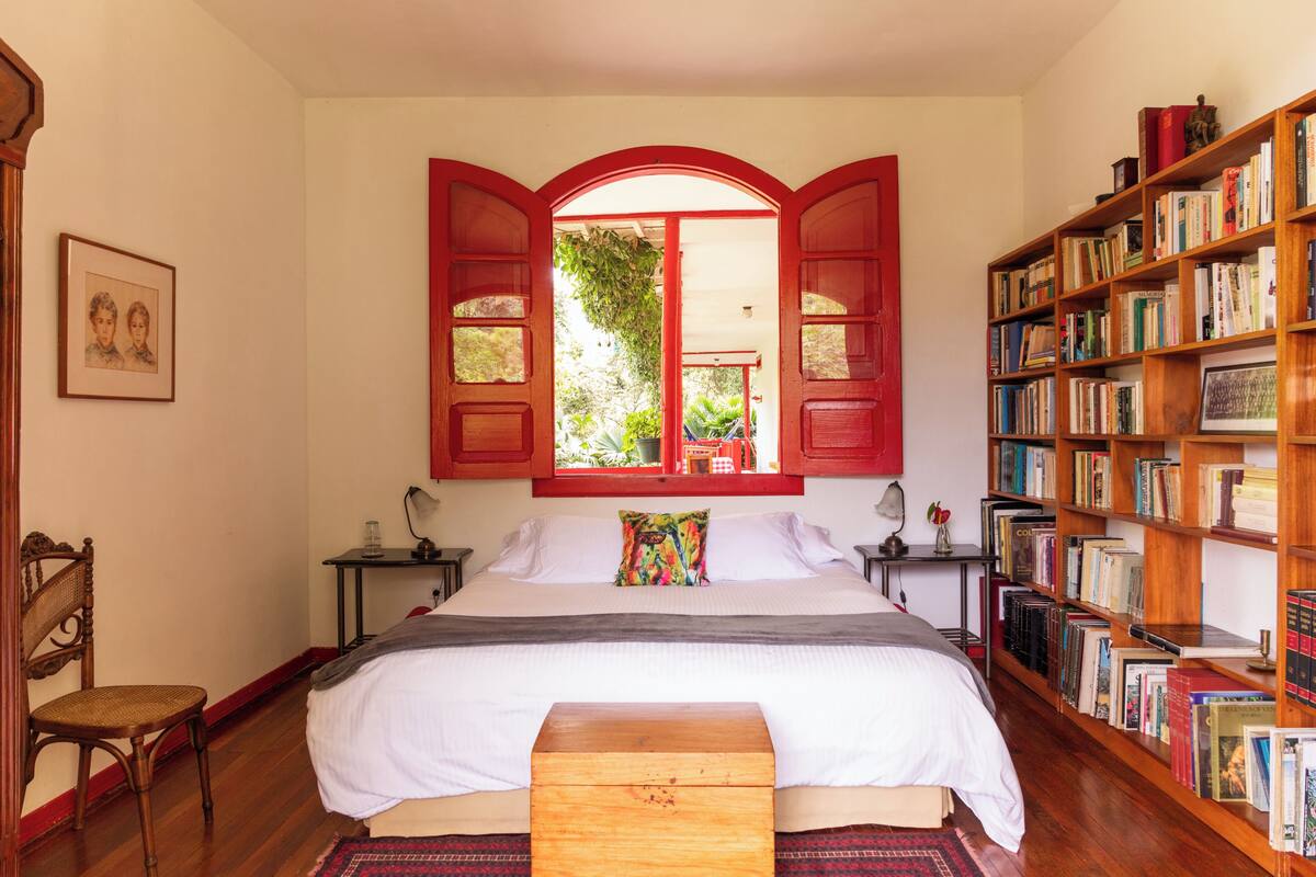 Un dormitorio con una cama grande, un banco de madera, una estantería y una ventana con persianas abiertas.
