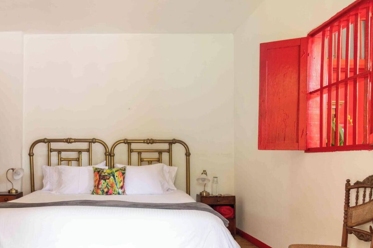 Una cama con estructura metálica, ropa de cama blanca y almohada colorida. Hay una mesita de noche con una lámpara y una pequeña mesa con un jarrón. Se ve una puerta roja y un armario rojo.