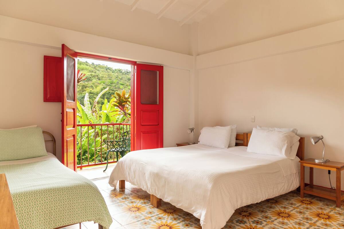 Un dormitorio con cama, mesitas de noche, ventana con vistas a zonas verdes y puerta al balcón.