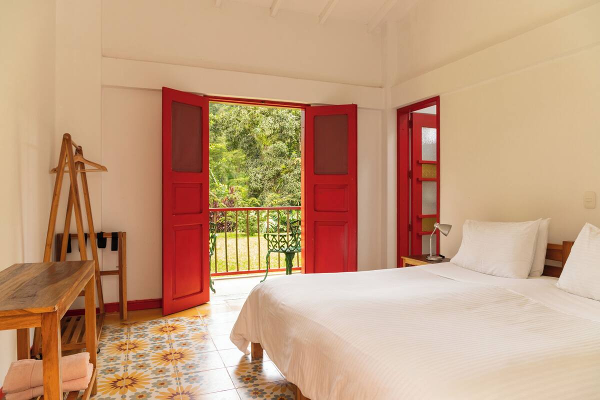 Un dormitorio con cama, una mesa de madera, una silla colgante y un balcón con puertas rojas.