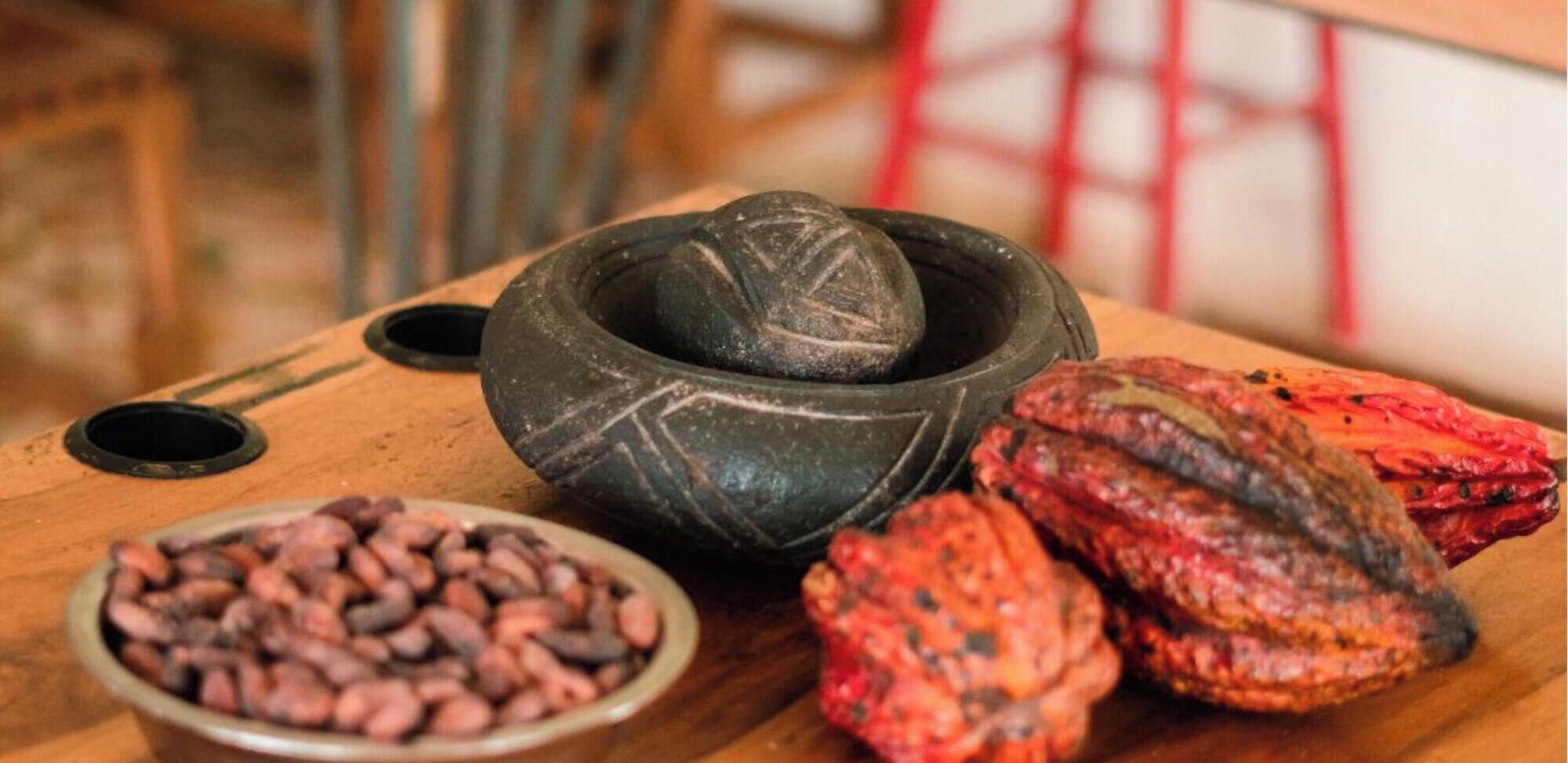 A wooden surface with a stone mortar, a bowl of beans, and roasted peppers.