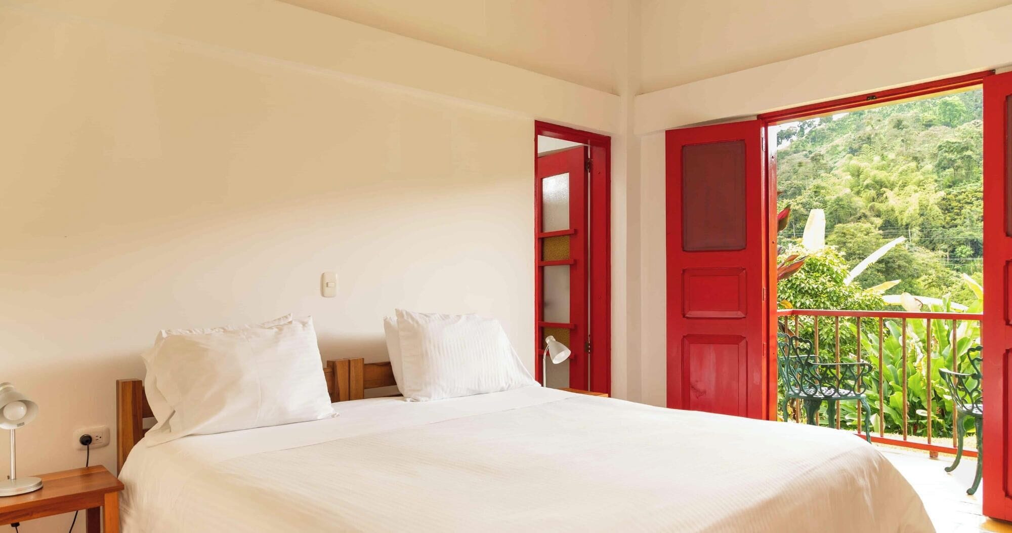 Un dormitorio con cama, mesita de noche, lámpara y una puerta roja que da a un balcón con vegetación.