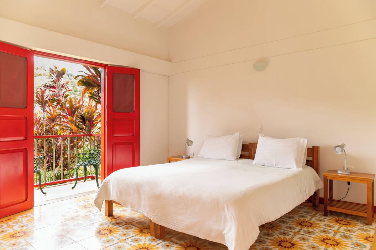 Un dormitorio con cama, dos mesitas de noche, un buró, una puerta roja y un balcón con plantas.