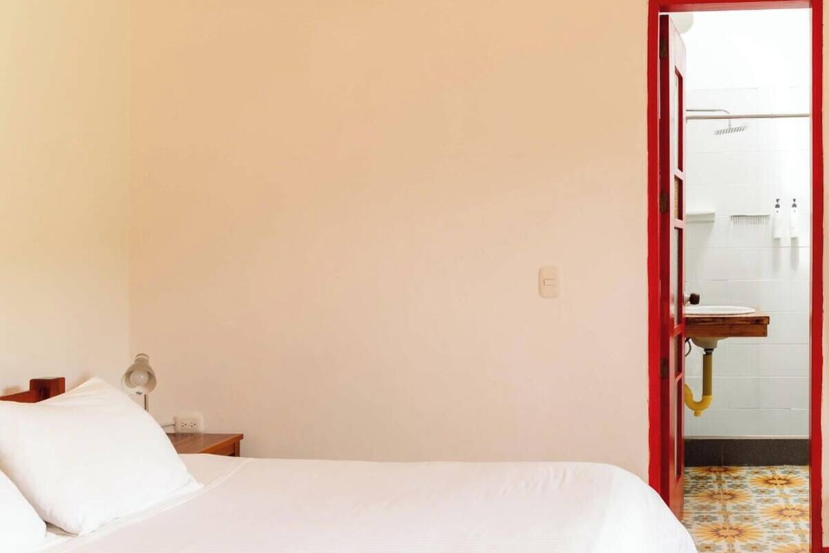 Un dormitorio con una cama, una mesita de noche, una lámpara y un baño visible a través de una puerta abierta.