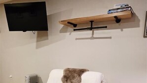 Una habitación con una silla de madera, una mesita pequeña, un televisor montado y una estantería con libros.