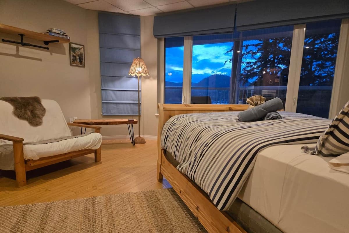 Ett sovrum med en säng, en stol, ett sängbord, en lampa och ett fönster med utsikt över träd.