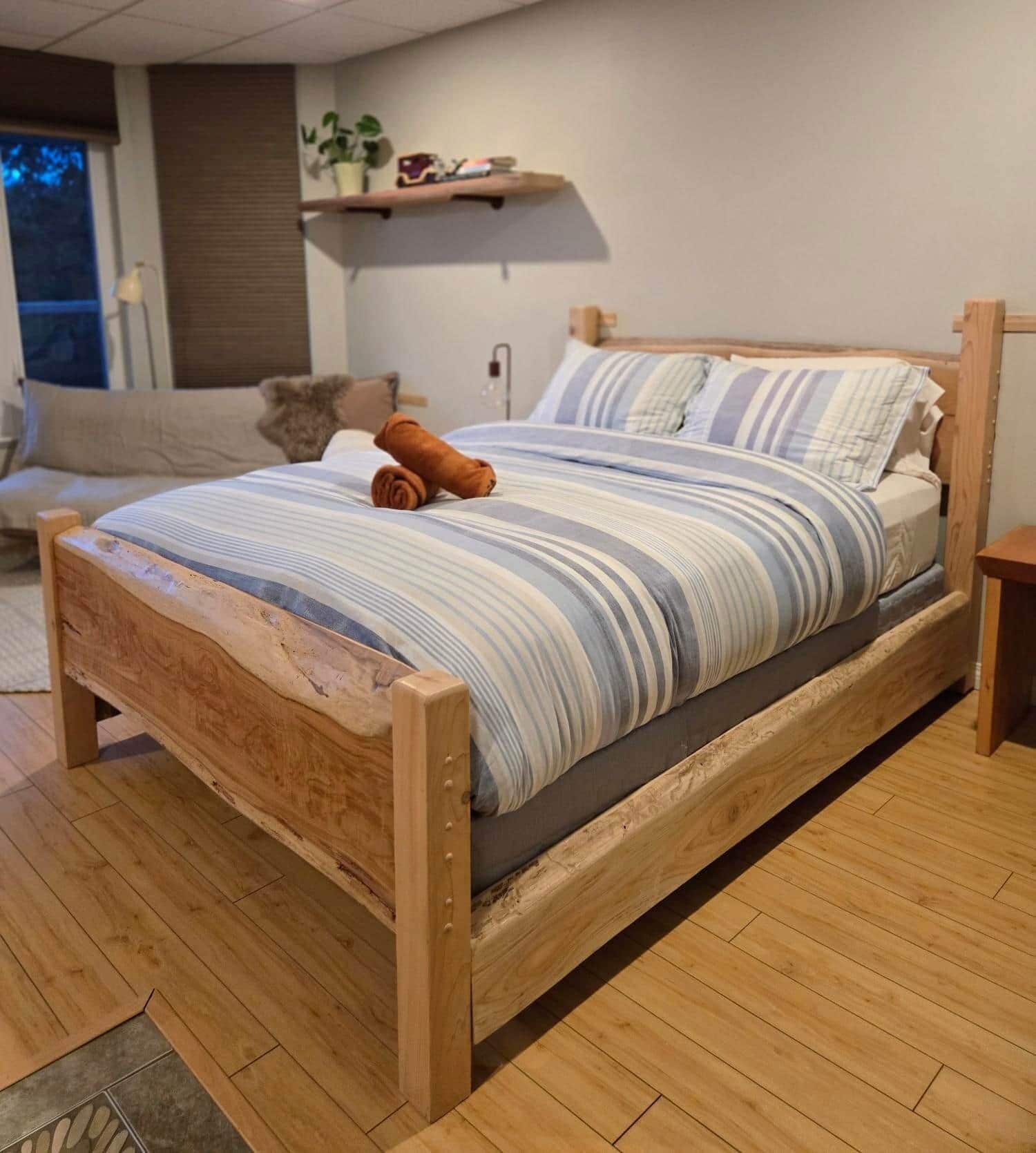 A wooden bed with a striped bedspread and pillows, a rolled towel, and a bedside table.