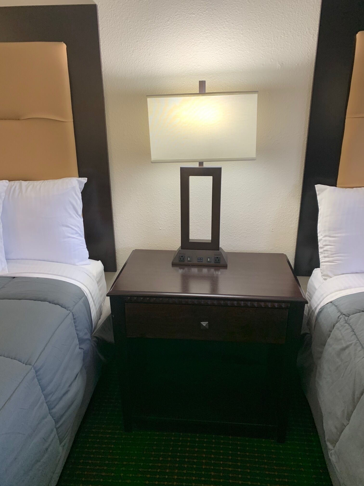 Une chambre d’hôtel avec deux lits simples, une table de chevet avec une lampe et un petit tiroir.
