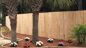A landscaped area with a wooden fence, two palm trees, and a variety of flowering plants. - Blackwater Inn (Milton)