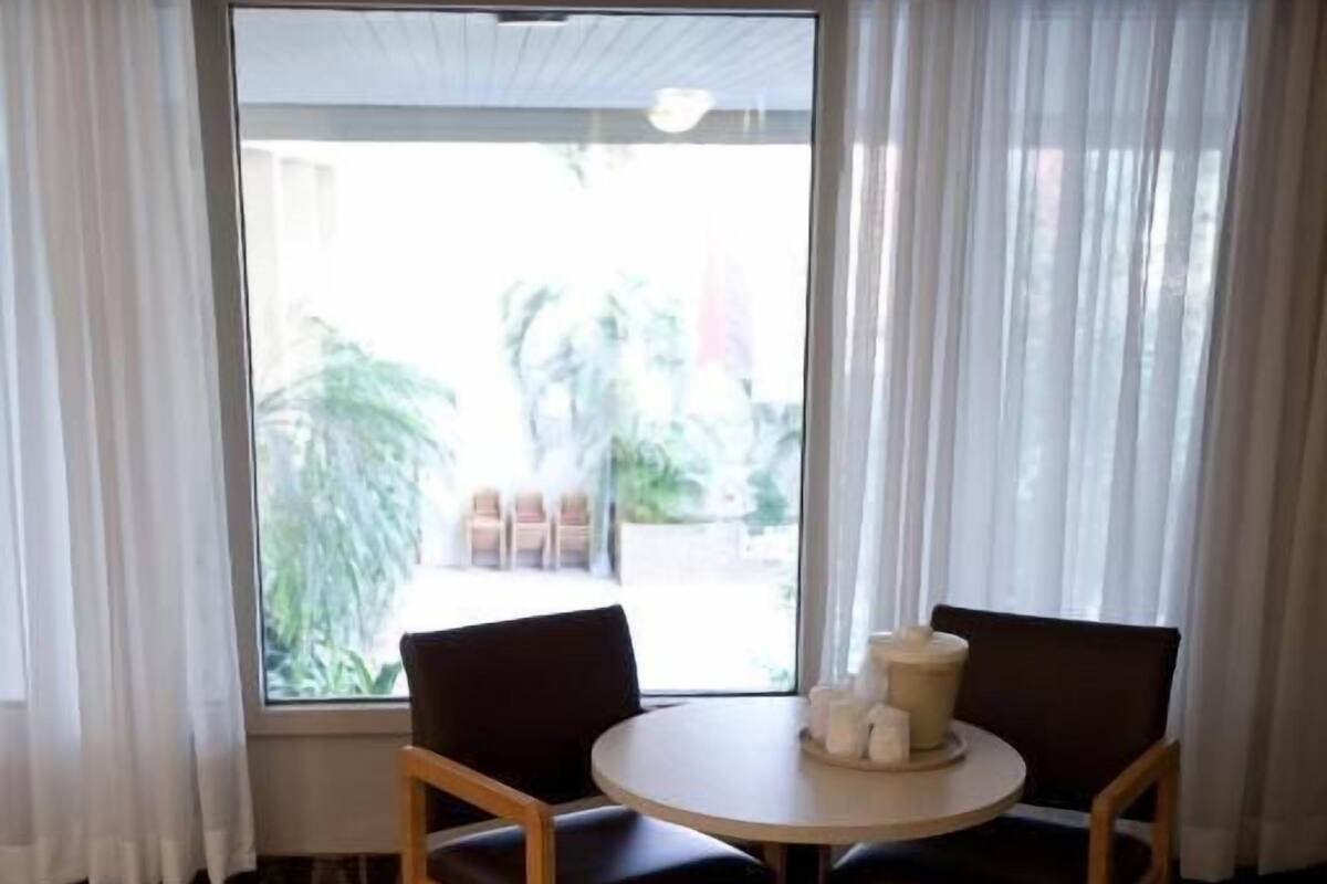 Quarto de hotel com cama, uma mesa redonda com duas cadeiras, uma janela com cortinas e piso coberto por carpete.