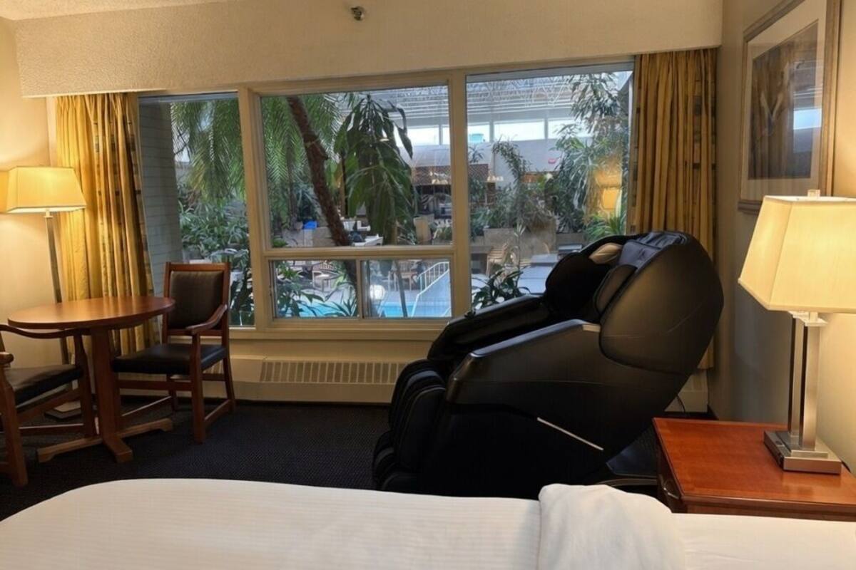 Quarto de hotel com cadeira de massagem, cama, escrivaninha e uma grande janela com vista para palmeiras e piscina.