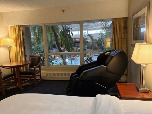 Habitación de hotel con sillón de masaje, cama, escritorio y ventana grande con vistas a palmeras y una piscina.