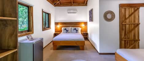 Un dormitorio con techo de madera, una cama con almohadas estampadas, un refrigerador pequeño y una ventana con vista a árboles.