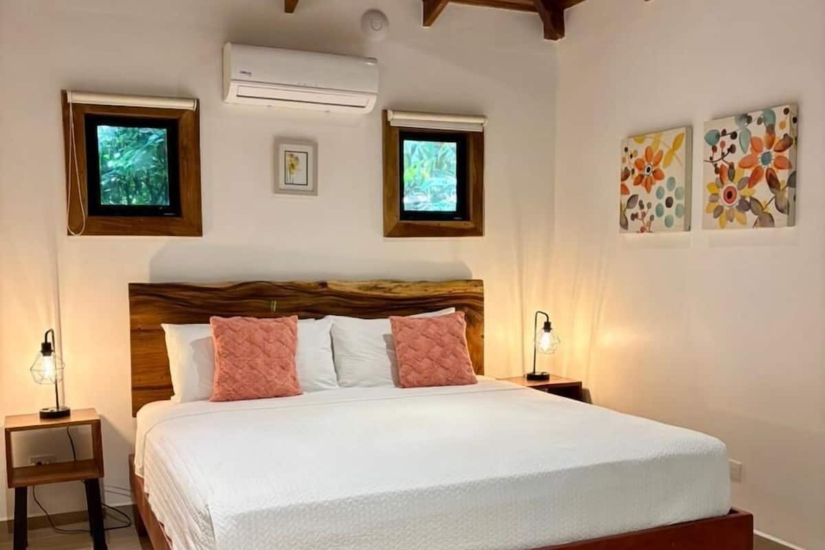 Ein Schlafzimmer mit Holzbalkendecke, einem großen Bett mit weißer Bettwäsche und pinken Kissen, zwei Wand-Fernsehern und zwei gerahmten Kunstwerken.