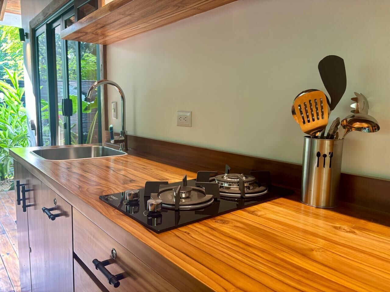 A modern kitchen with a wooden countertop, stainless steel appliances, and a large window overlooking greenery.