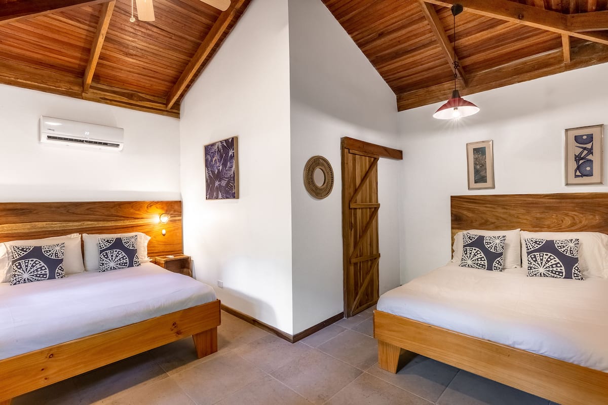 Ein Zimmer mit zwei Holzbett, weißen Wänden, Holzbalken an der Decke und einem Deckenventilator.