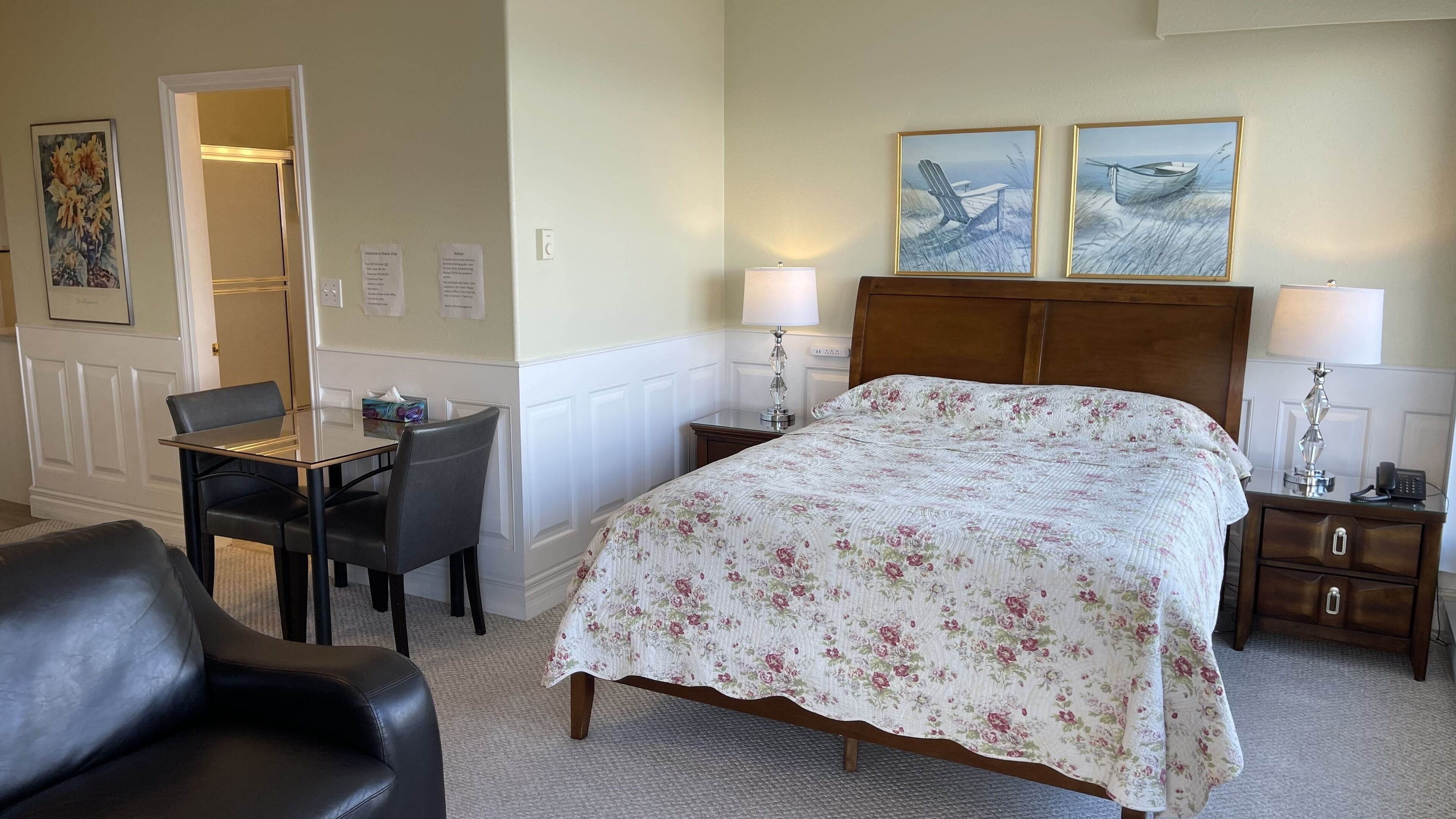 Habitación de hotel con cama, dos lámparas de noche, una mesita de noche de madera, un escritorio con silla y una puerta que da a otra habitación.