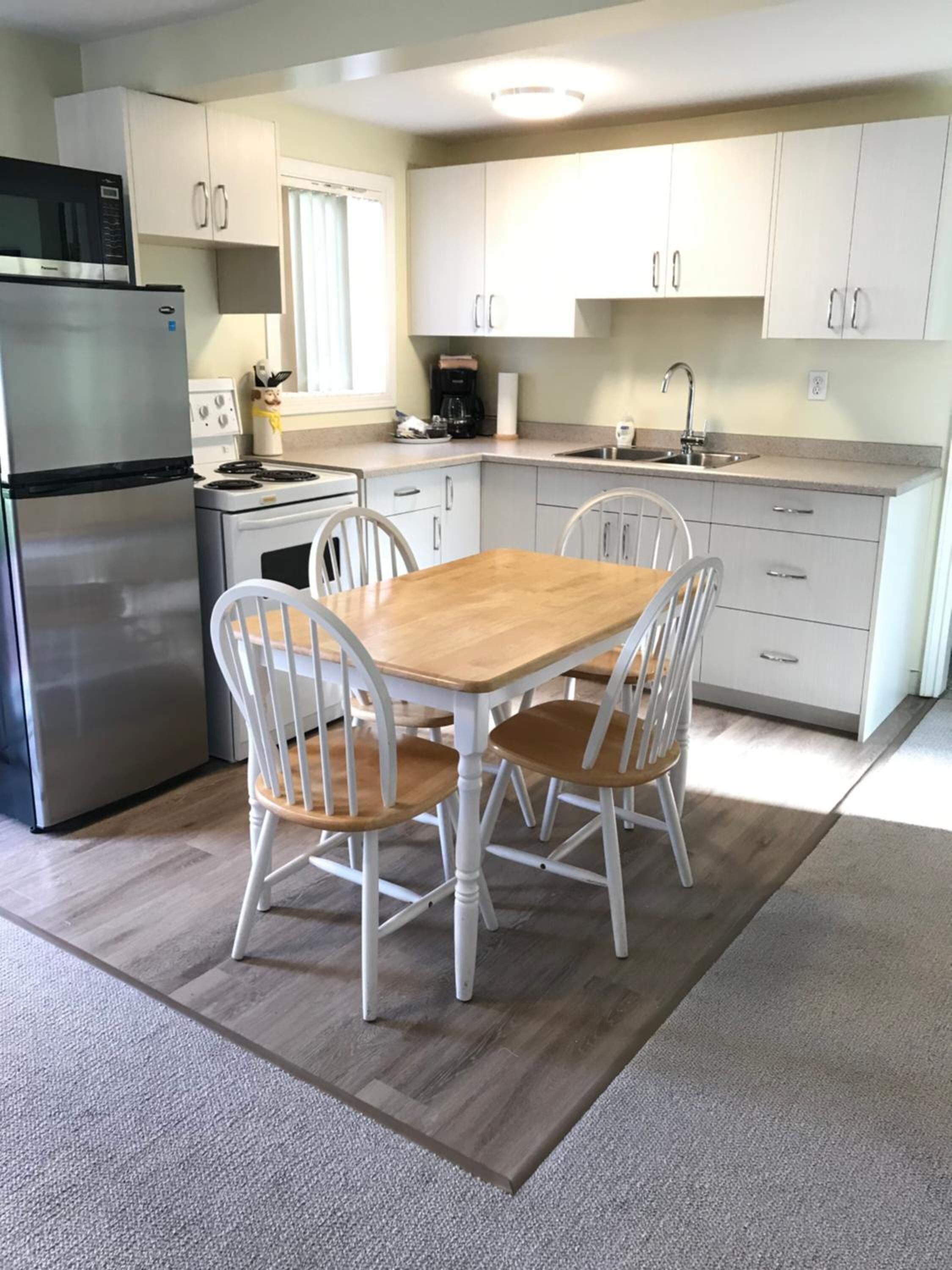 Una cocina compacta con armarios blancos, un refrigerador de acero inoxidable, un microondas, una estufa y un fregadero. Hay una mesa de madera con cuatro sillas.