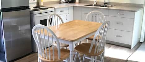 A compact kitchen with white cabinets, a stainless steel refrigerator, a microwave, a stove, and a sink. There is a wooden table with four chairs.