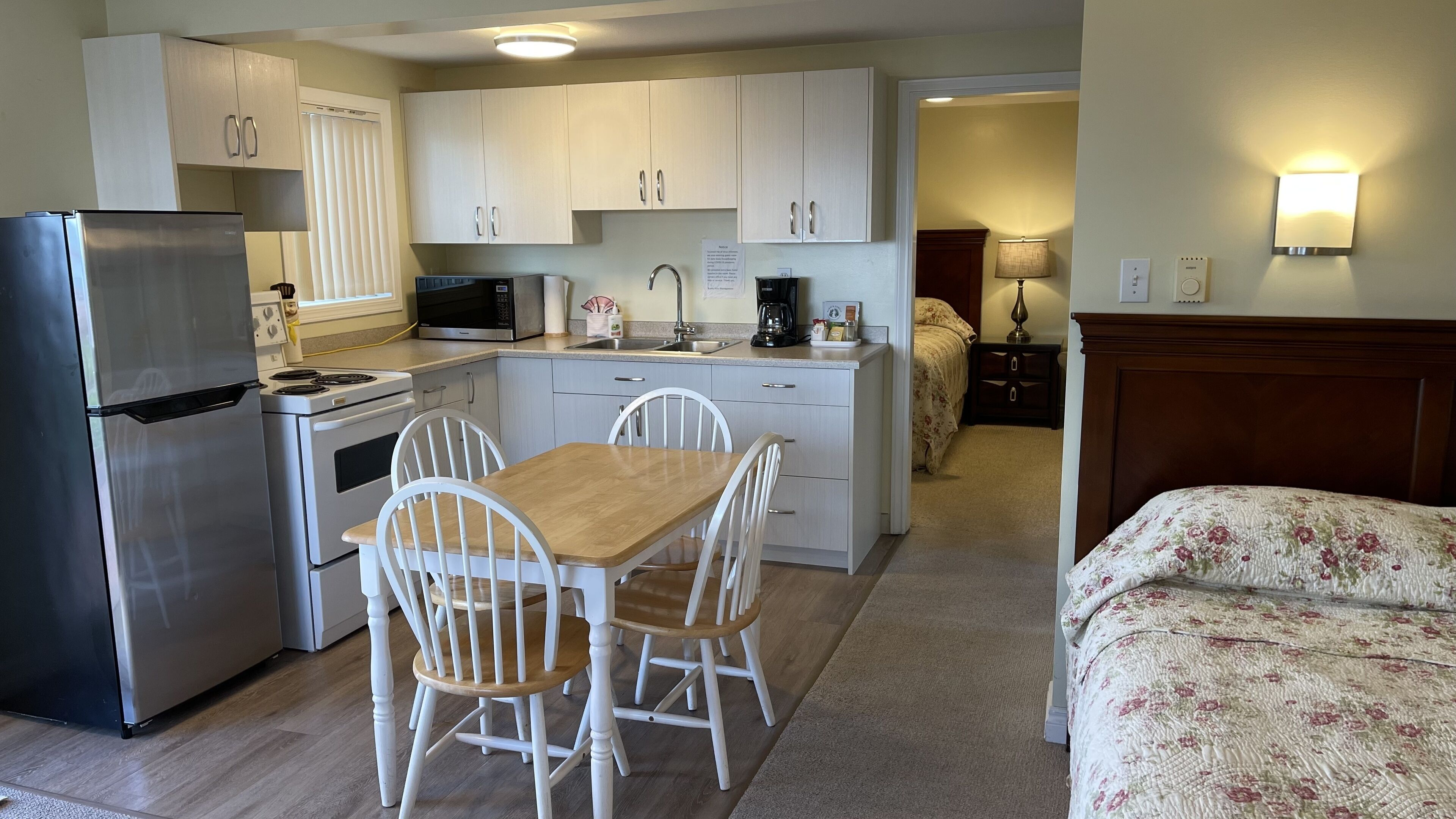 A compact kitchen with white cabinets, a refrigerator, stove, microwave, and a dining area with a wooden table and white chairs.