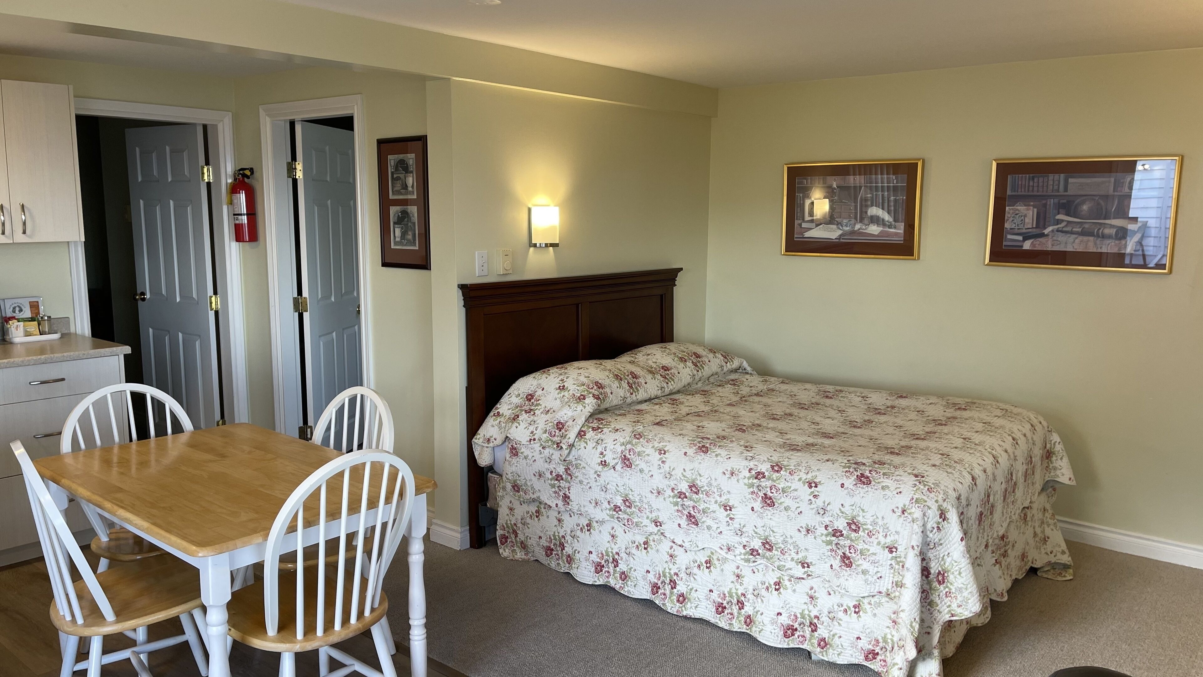 A room with a wooden table and white chairs, a bed with floral bedding, and two framed pictures on the wall.