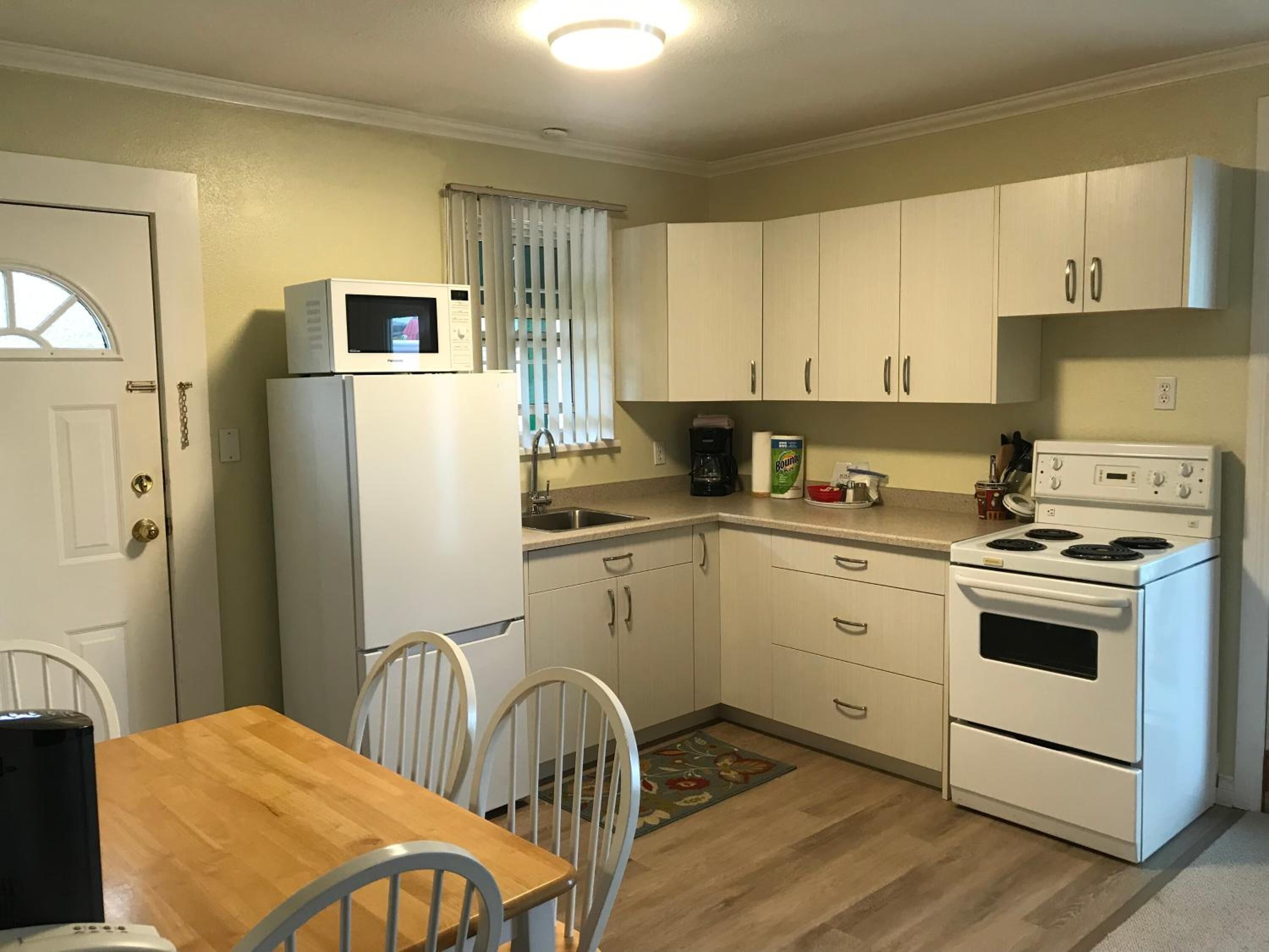 Cocina compacta con armarios blancos, microondas, refrigerador, estufa y cafetera. Hay una mesa de madera con sillas y una ventana con persianas.